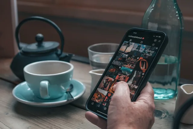 A person holds a smartphone browsing Instagram next to a teacup and teapot on a wooden table. How to set up Instagram Shopping UK for your business