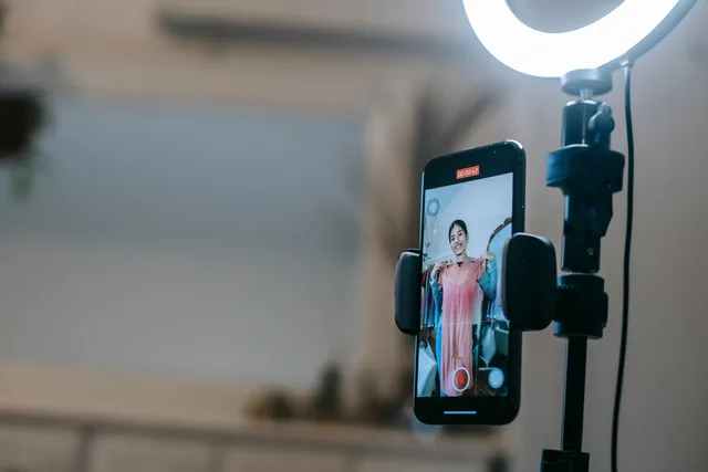 Anonymous female blogger standing in room and shooting vlog on smartphone with ring lamp while demonstrating pink dress in hands. Five English social commerce agencies by service boundary, with no ranking implied
