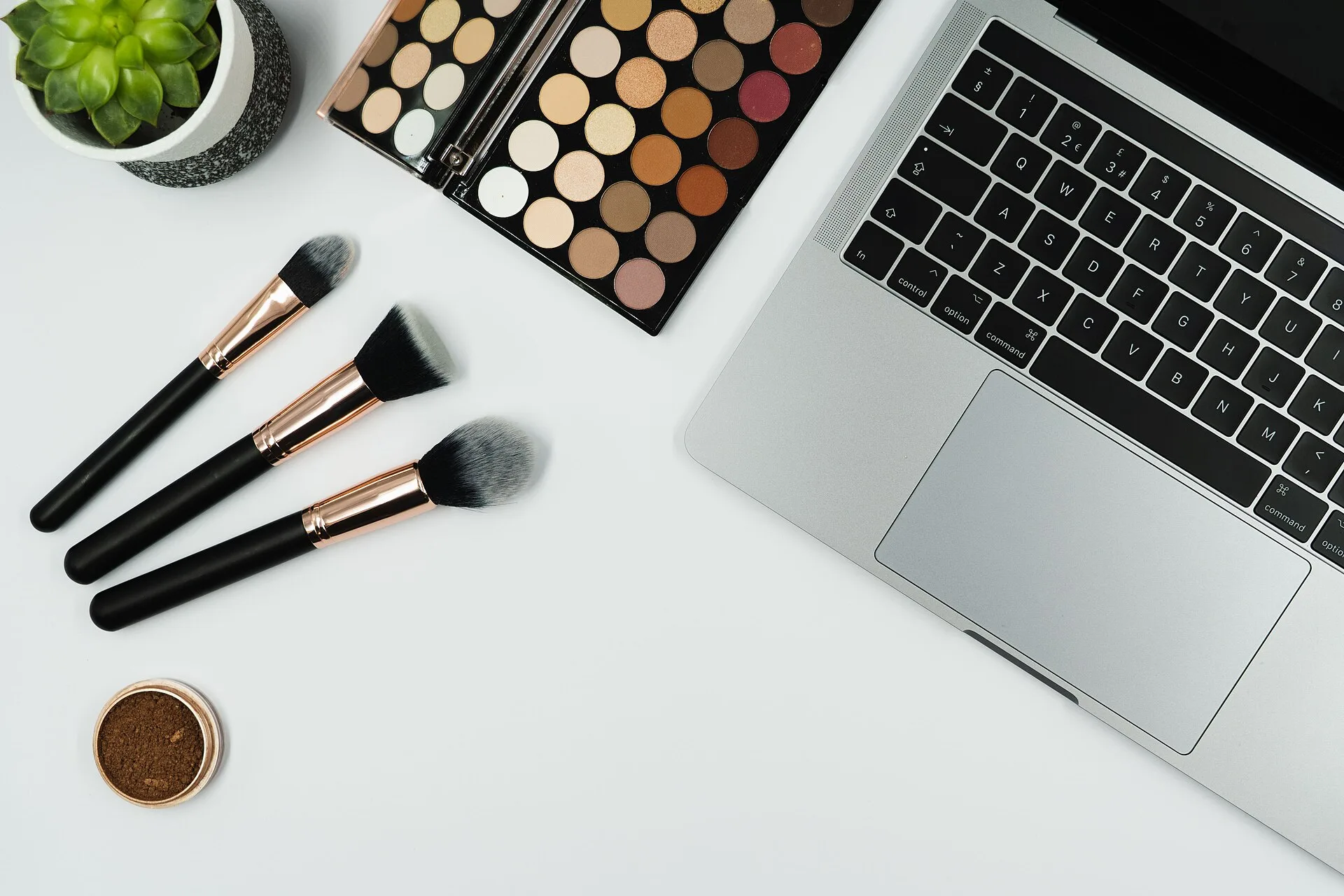 In this photo a person's desk can be seen whilst they get ready for a zoom call. Make up and brushes are pictured, next to a laptop on a white background. We initially took this image for a blogpost we planned on the topic of putting on makeup for a zoom call, but have now chosen to release it under
