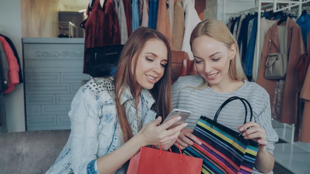 Two women enjoying a shopping spree and sharing a moment on their smartphone in a clothing store. Social commerce in England for 2027 starts by defining the transaction, not the market