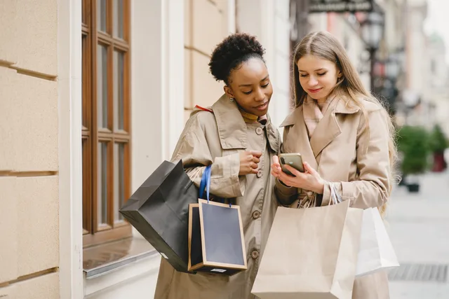 Two women with shopping bags check their phone on a city street. Why IDBR cannot show social commerce market size in England