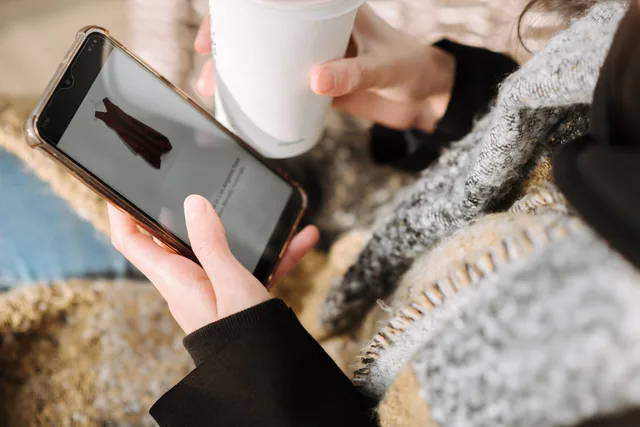 From above of crop anonymous female purchaser with takeaway hot drink choosing dress on cellphone screen during online shopping. Social commerce returns policy UK: refunds and deductions