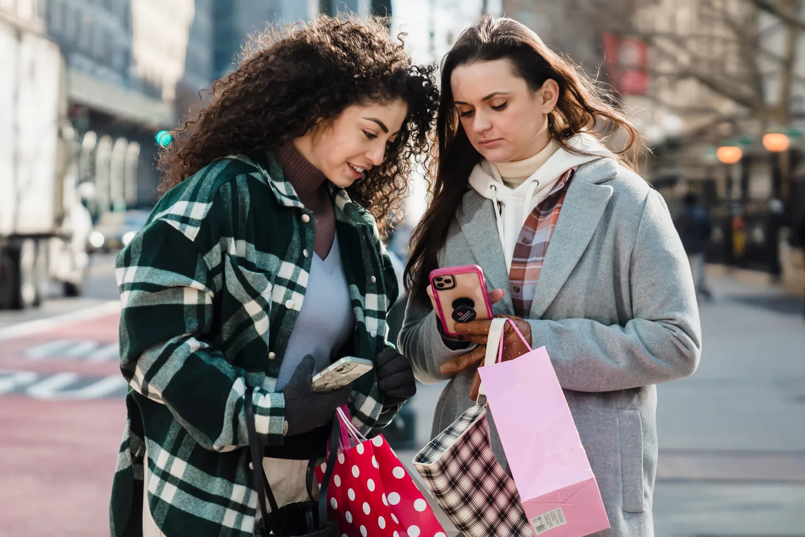 Calm women friends in trendy clothes standing with shopping bags on city street and browsing smartphone in sunny day