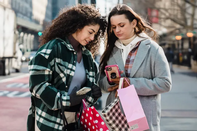 Calm women friends in trendy clothes standing with shopping bags on city street and browsing smartphone in sunny day. Budgeting before checking eligibility, and other social commerce planning mistakes
